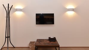 A room with a flat-screen TV mounted on the wall, a wooden table with a basket, a trash bin, and a floor lamp. - COCOSHUKU Tokyo Nakano (Tokyo)