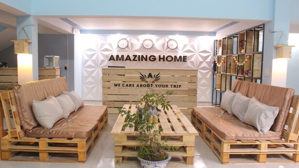 A reception area with a wooden bench seating arrangement, a potted plant, and a wall with the text "AMAZING HOME WE CARE ABOUT YOUR TRIP". - Ha Giang Amazing Hostel (Ha Giang 2)
