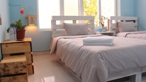 A bedroom with two beds, wooden headboards, a bedside table with a lamp, a potted plant, and a window with a view of trees. - Ha Giang Amazing Hostel (Ha Giang 2)