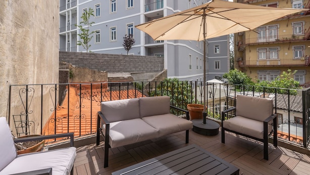 A balcony with a beige sofa, a small round table, and an umbrella. The balcony overlooks a red-tiled roof and a building with multiple balconies.