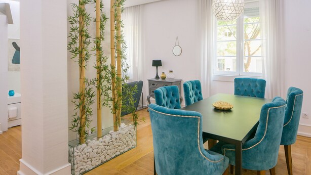 Un espace repas avec une table verte et des chaises recouvertes d’un tissu bleu, une cloison en verre décorée de petits galets blancs et des plantes en bambou.
