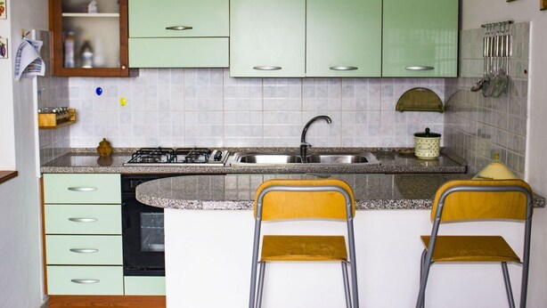 Een compacte keuken met groene kastjes, een granieten aanrecht, een spoelbak en twee gele stoelen.