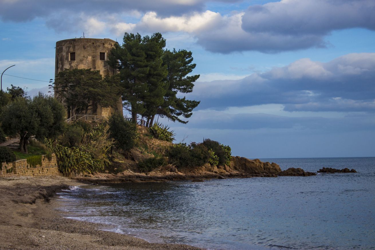 A coastal scene with a historical stone tower, rocky shoreline, and dense greenery.