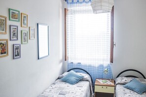 Et soveværelse med to enkeltsenge, et natbord med lampe, et vindue med ternet gardin og flere billeder i rammer på væggen.