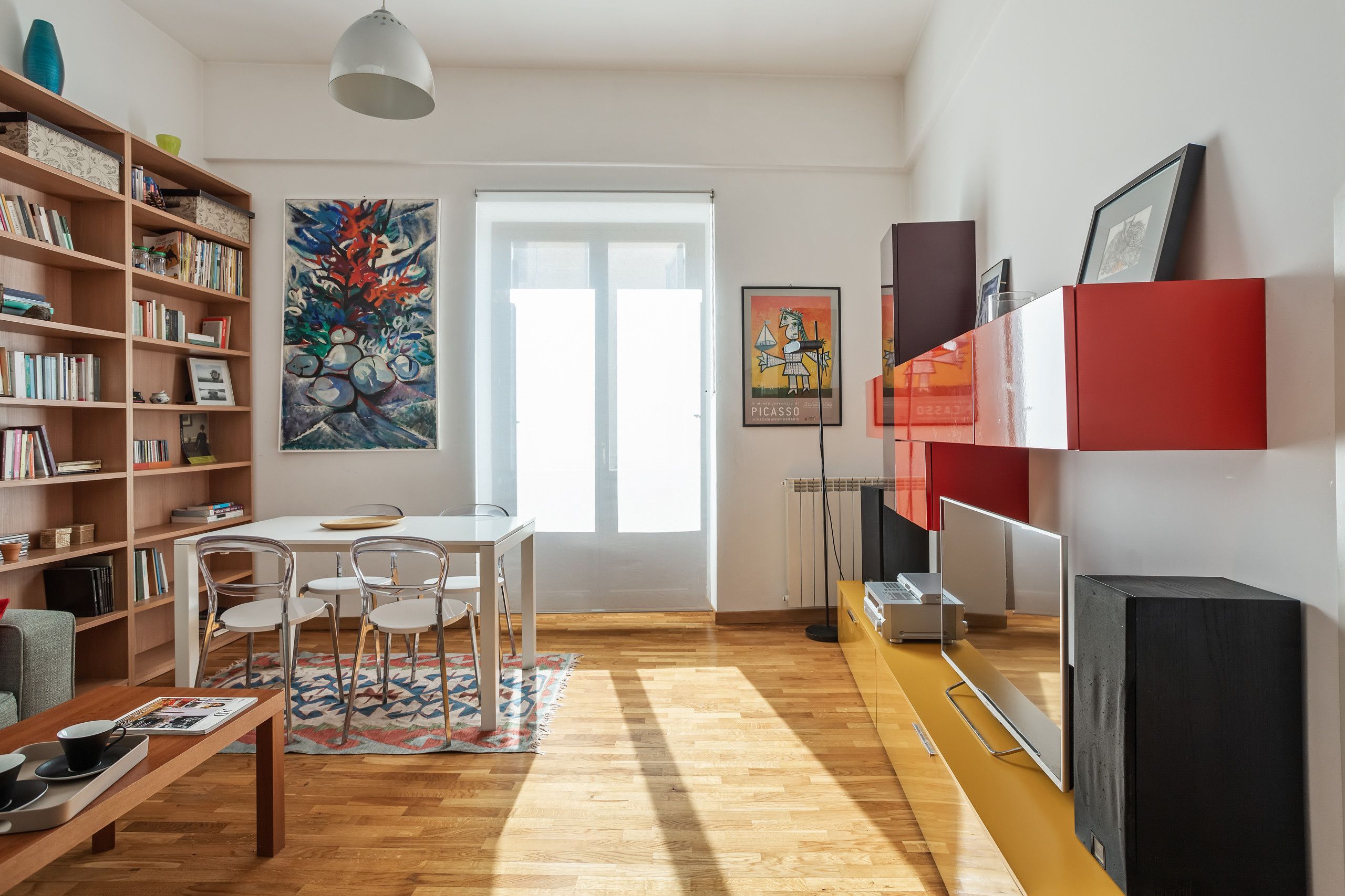A modern living room with a wooden floor, a dining area with a table and chairs, a bookshelf, a painting, and a television.