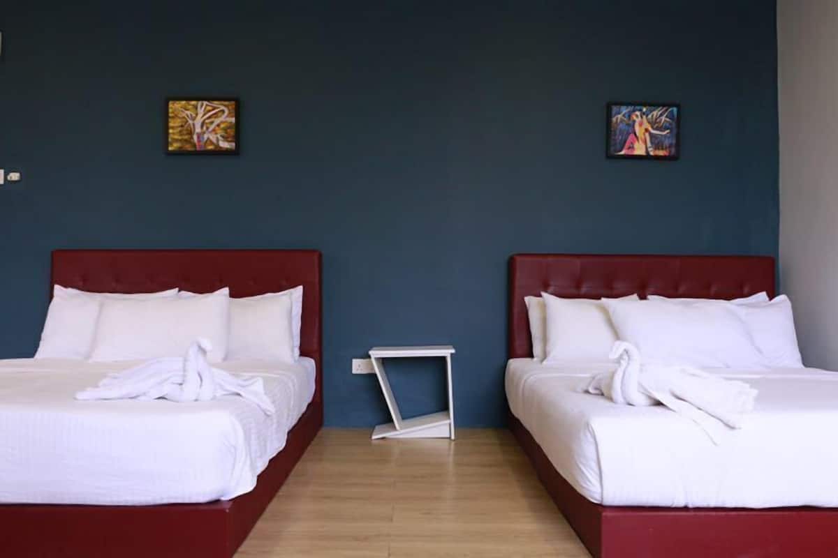 Quarto de hotel com duas camas, cada uma com roupa de cama branca e cabeceiras vermelhas. Há duas pinturas na parede, uma pequena mesa de cabeceira branca e piso de madeira.