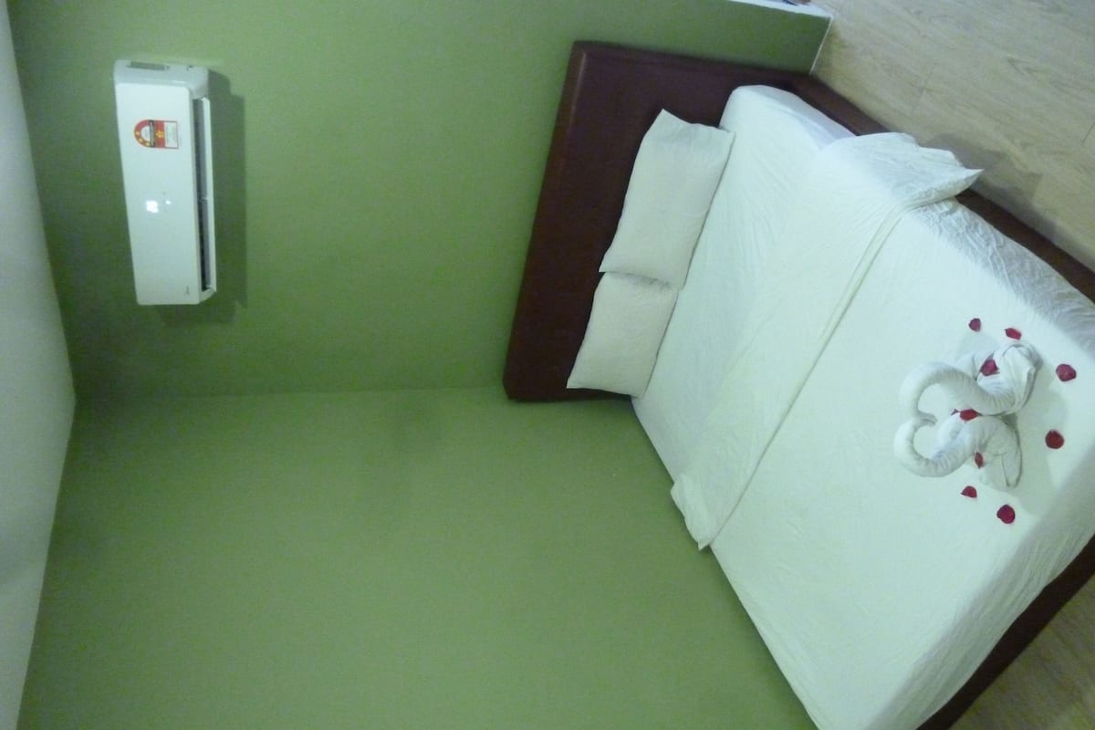Quarto de hotel com chão verde, unidade de ar condicionado branca, cama com roupa de cama branca e travesseiro decorativo em forma de cisne, e cabeceira marrom.