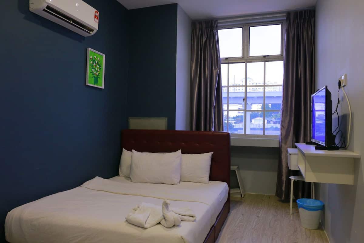 Quarto de hotel com cama, escrivaninha com computador, janela com cortinas e ar condicionado.