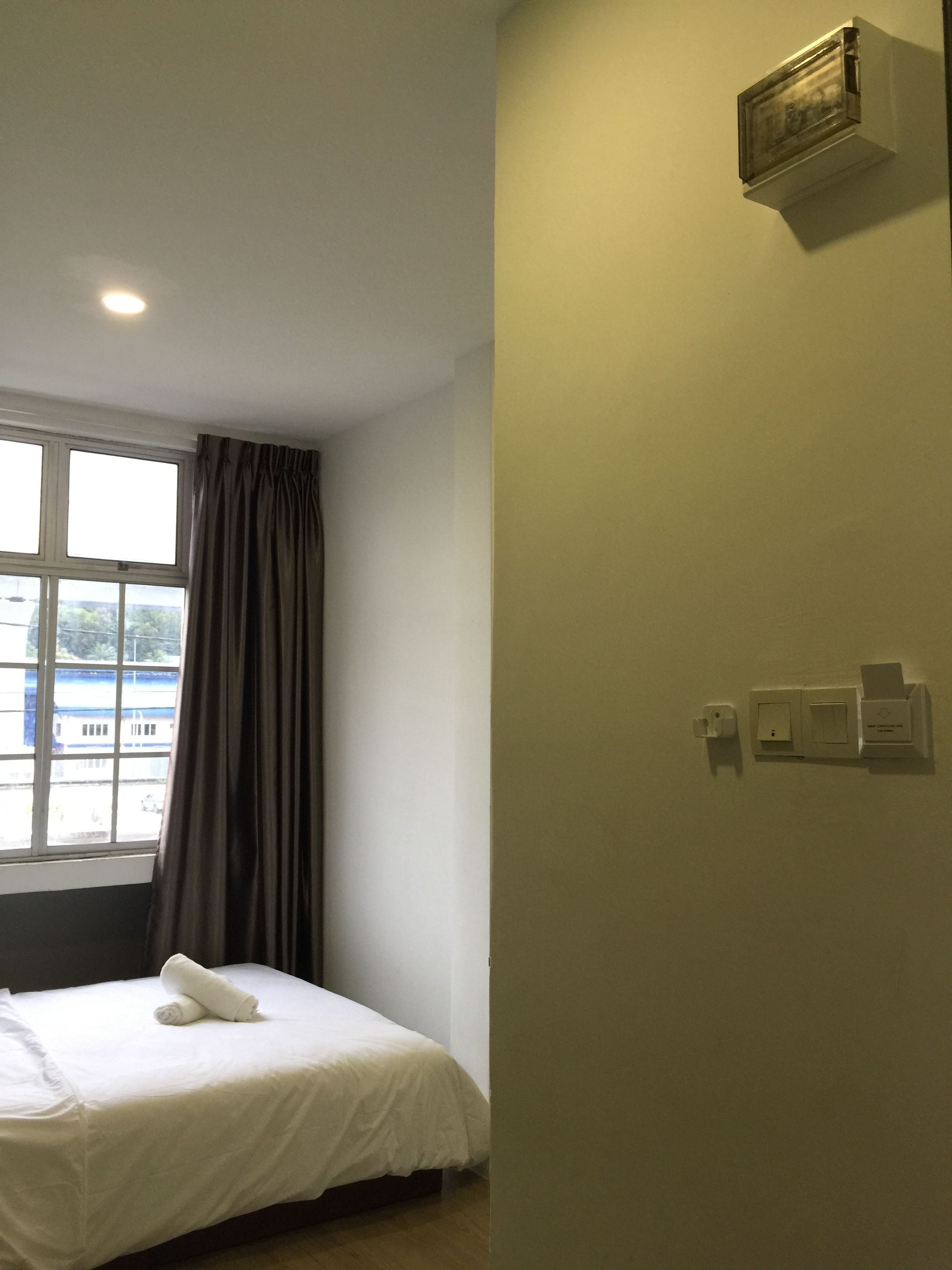 A hotel room with a bed, a window with curtains, a wall-mounted air conditioning unit, and a wall socket.