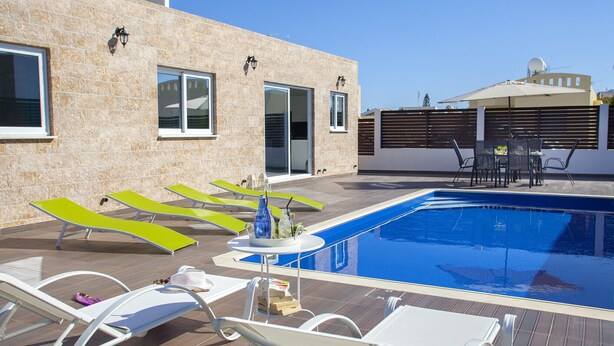 A swimming pool with lounge chairs, a table with drinks, and a building with a door and windows. - Protaras Villa Hollywood Near To The Centre (Protaras)