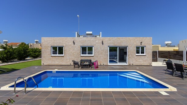 Una piscina rectangular con plataforma empapelada, un edificio de techo plano y un cielo azul despejado.