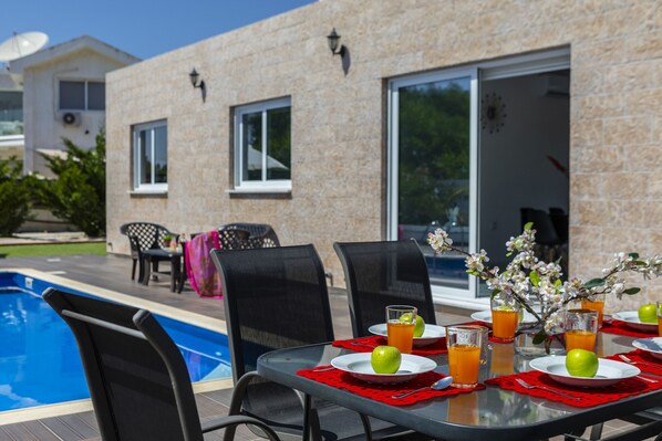 An outdoor dining area with a pool, chairs, and a table set with plates, glasses, and a vase of flowers. - Protaras Villa Golden Dream Near To The Centre (Protaras)