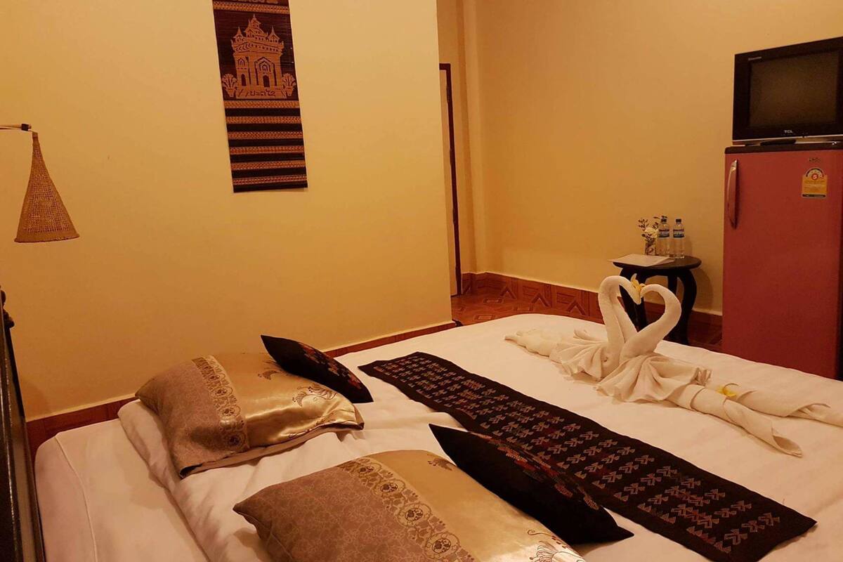 A hotel room with a bed, pillows, a towel, a keyboard, a small table with a vase, a TV, and a refrigerator.