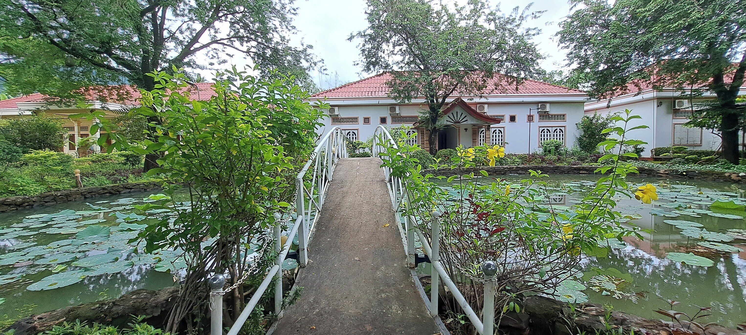 A bridge leading to a building with a red roof, surrounded by lush greenery and a pond with lily pads.