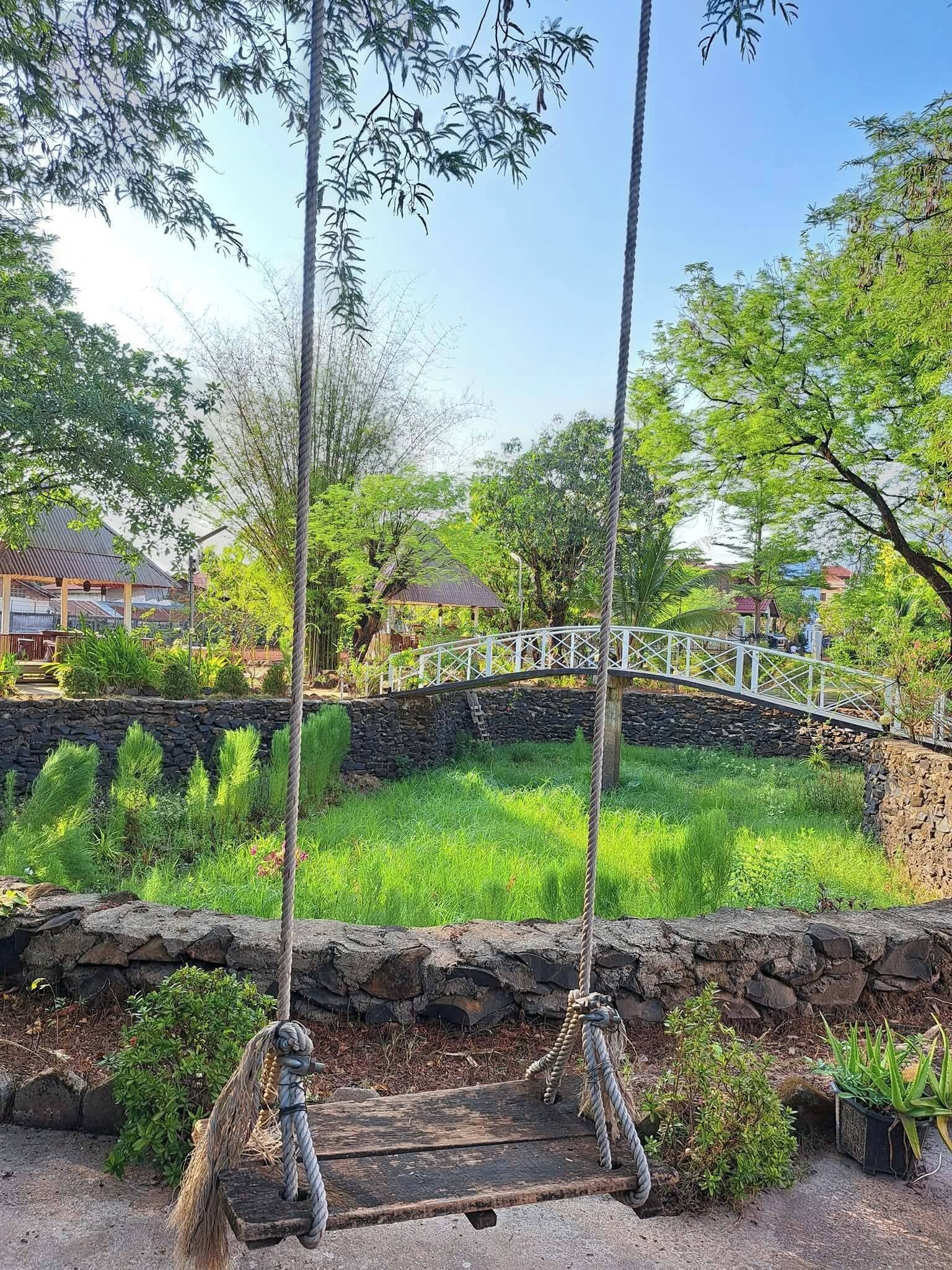 A wooden swing set with ropes, a stone wall, and a white bridge in a garden.