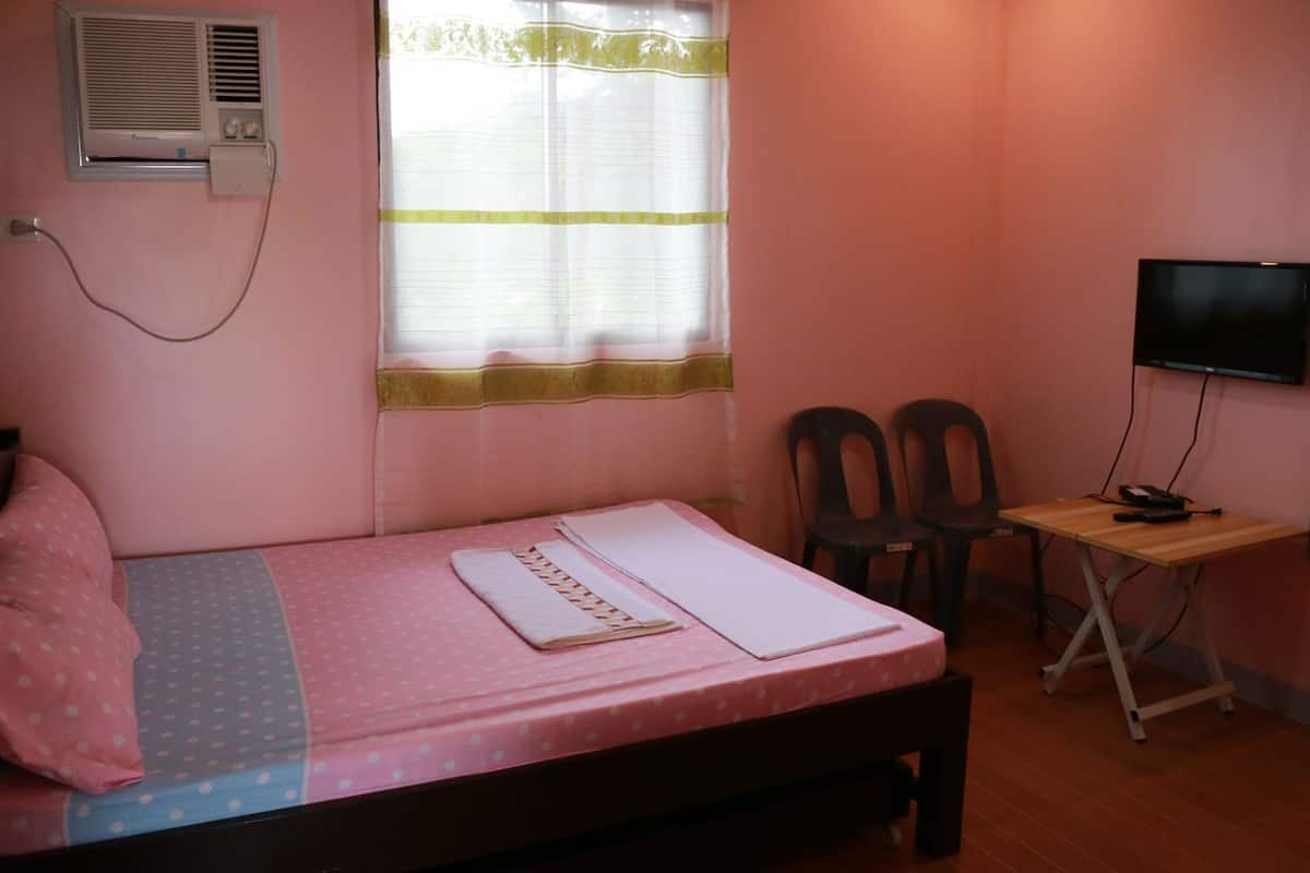 A single bed with a pink and blue polka dot bedspread, a small desk with a chair, a television, and an air conditioning unit mounted on the wall.