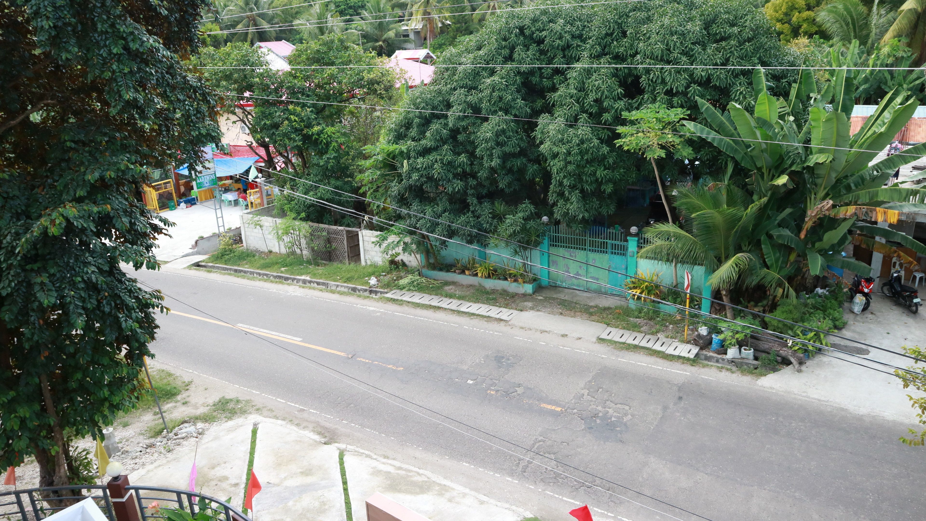A street view with a road, greenery, and a building with a blue fence.