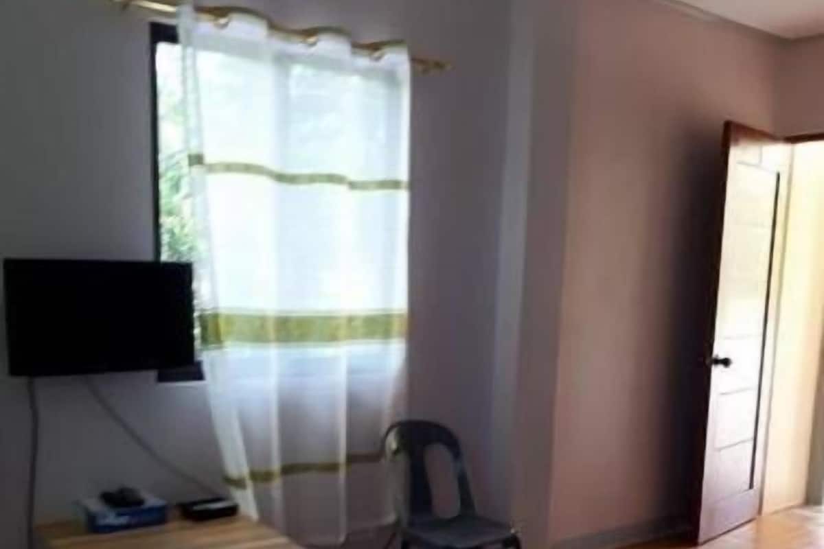 A room with a wooden desk, a chair, a television mounted on the wall, and a window with a curtain.