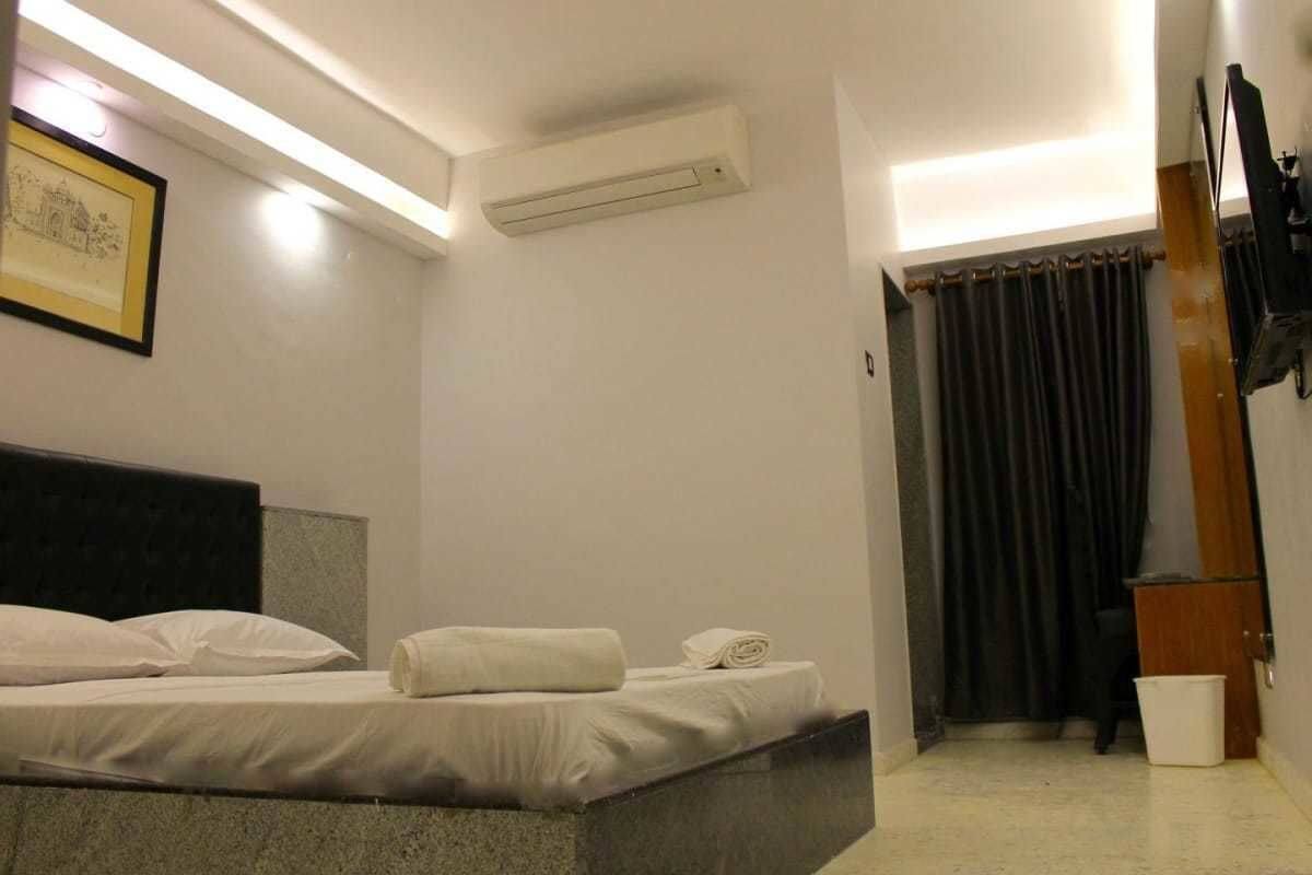 A hotel room with a bed, a wall-mounted air conditioner, a framed picture, and a curtain.