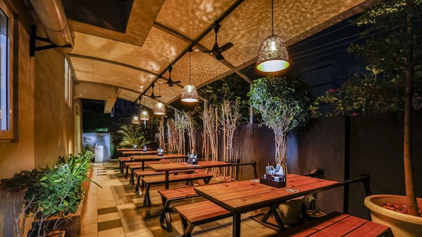 A row of wooden tables and benches under hanging lamps, with potted plants along the walls. - Floressa Randa (Chennai)