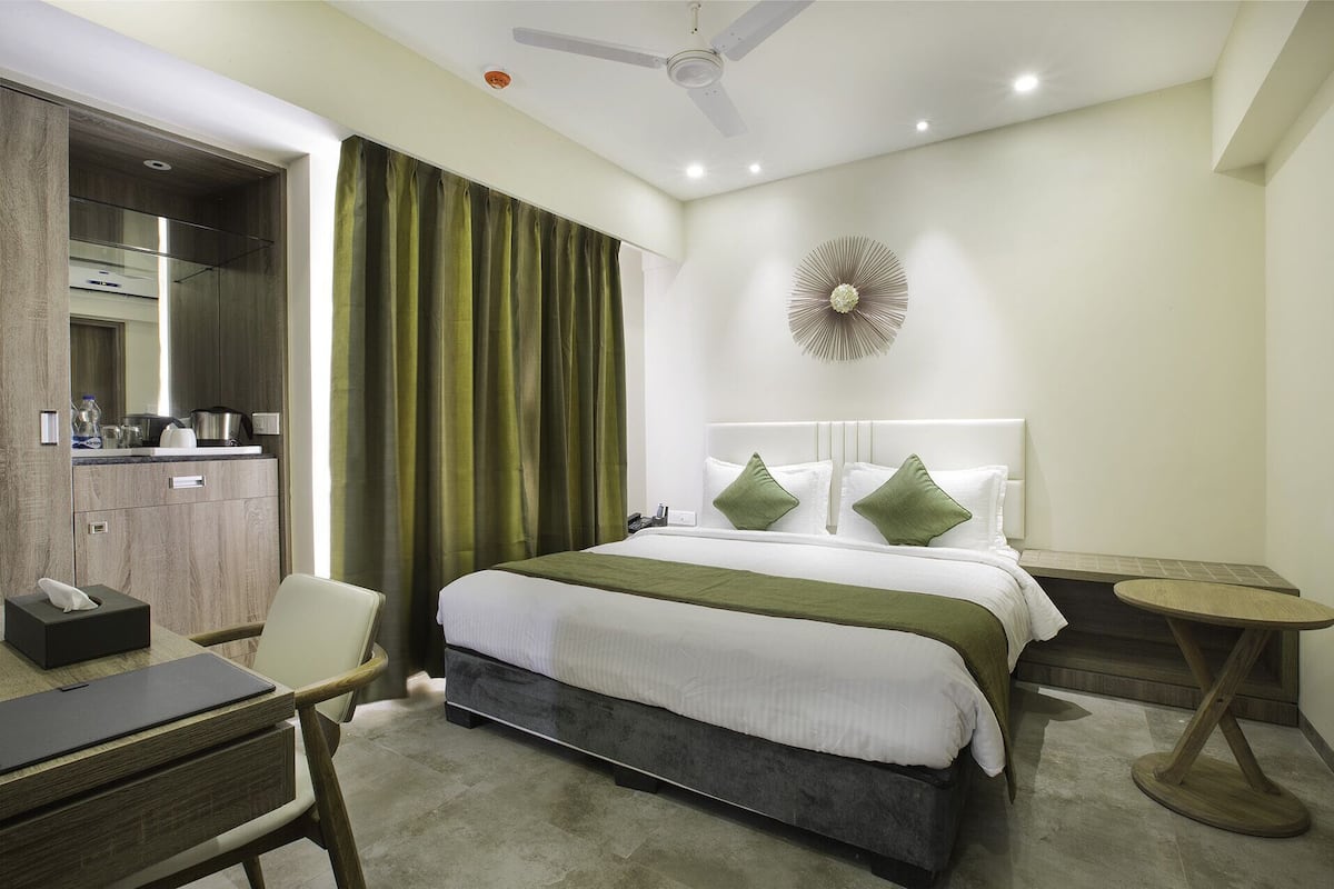 A hotel room with a bed, a desk, a chair, a small table, a ceiling fan, and a wall-mounted decorative piece.