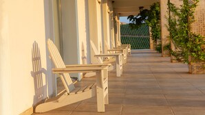 A row of white lounge chairs aligned against a building's exterior wall, with a concrete pathway leading to a garden area. - Villa Estet Bag Oteli (Kula)