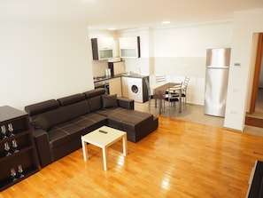 Una sala moderna con un sofá seccional, una mesita y un comedor con mesa y sillas. Se ve una cocina con lavadora y refrigerador al fondo.