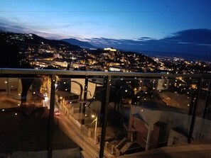 Vista nocturna desde un punto elevado que domina un paisaje urbano con edificios iluminados y una cadena montañosa distante.