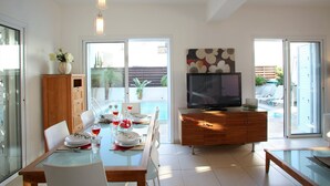 A modern dining area with a glass table, white chairs, and a wooden cabinet with a TV. - Villa Villa Kos 19 Near Konnos Beach (Protaras)