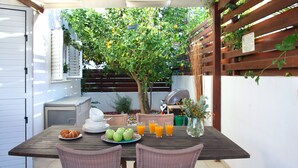 An outdoor patio with a wooden table set for two, surrounded by wicker chairs. There is a grill and a potted plant in the background. - Villa Villa Kos 19 Near Konnos Beach (Protaras)