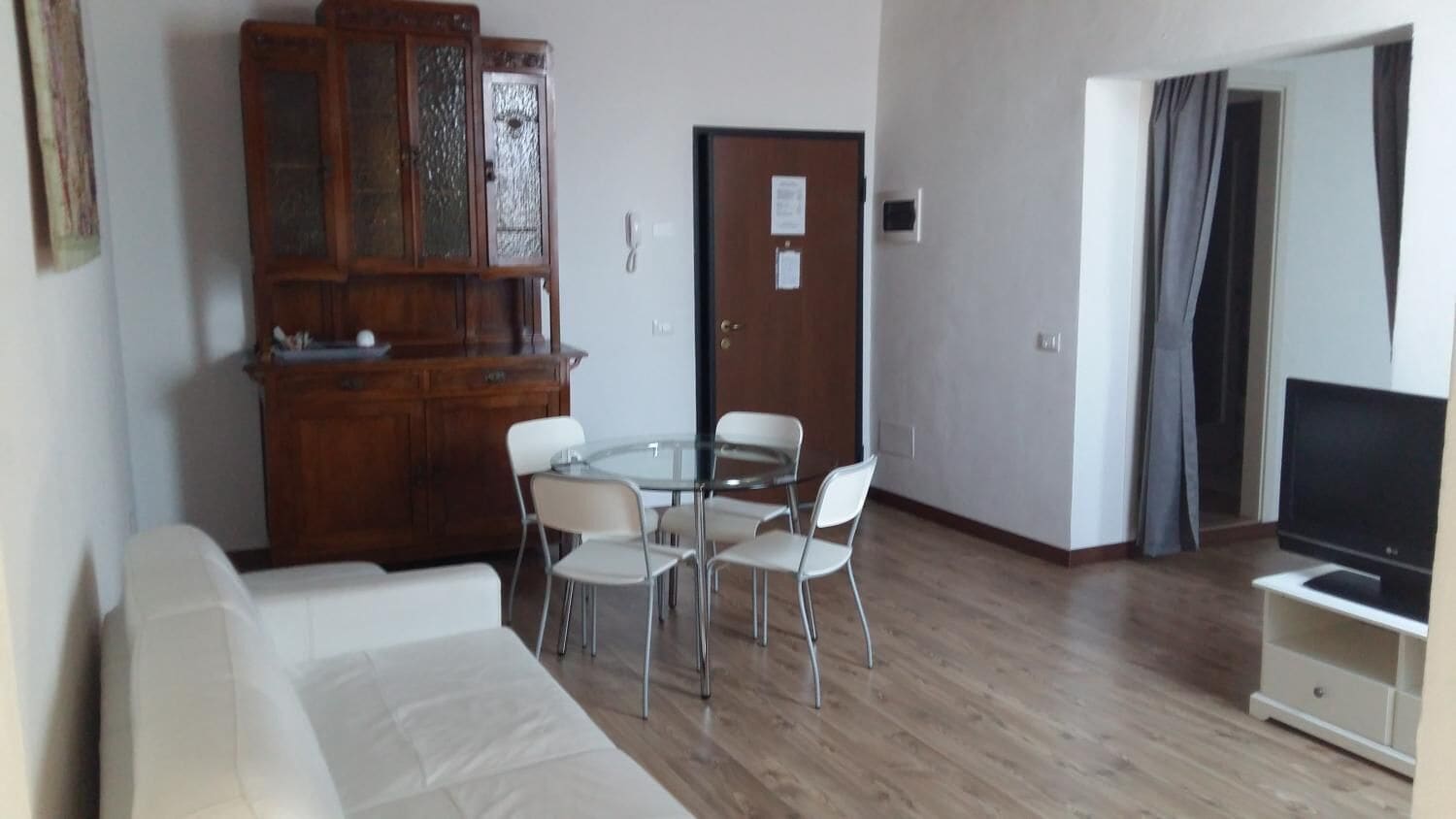 A living room with a white sofa, a glass-top dining table with four chairs, a wooden cabinet, and a TV on a stand.