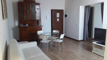 A living room with a white sofa, a glass-top dining table with four chairs, a wooden cabinet, and a TV on a stand.