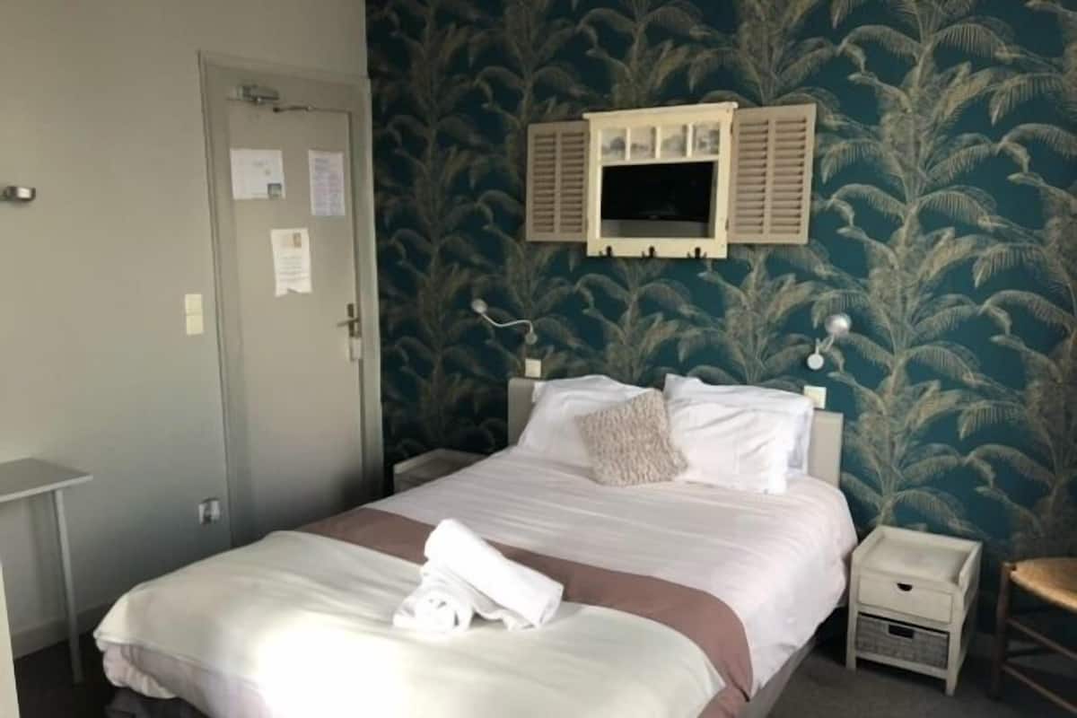 A neatly made bed with white linens and a brown accent, a bedside table with a lamp, and a wall-mounted TV.