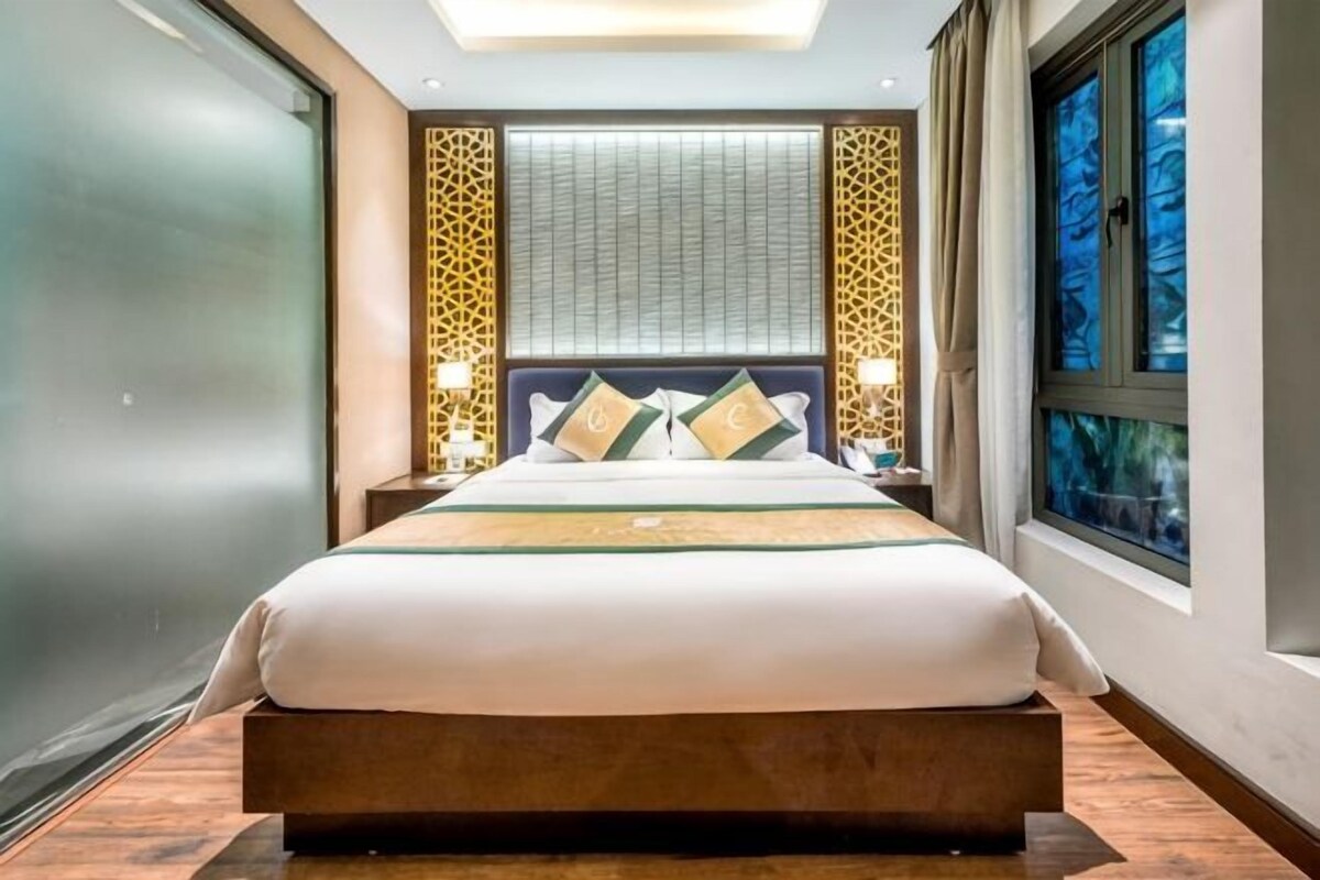 A hotel room with a large bed, a headboard with decorative panels, a window with a view, and a nightstand with a lamp.