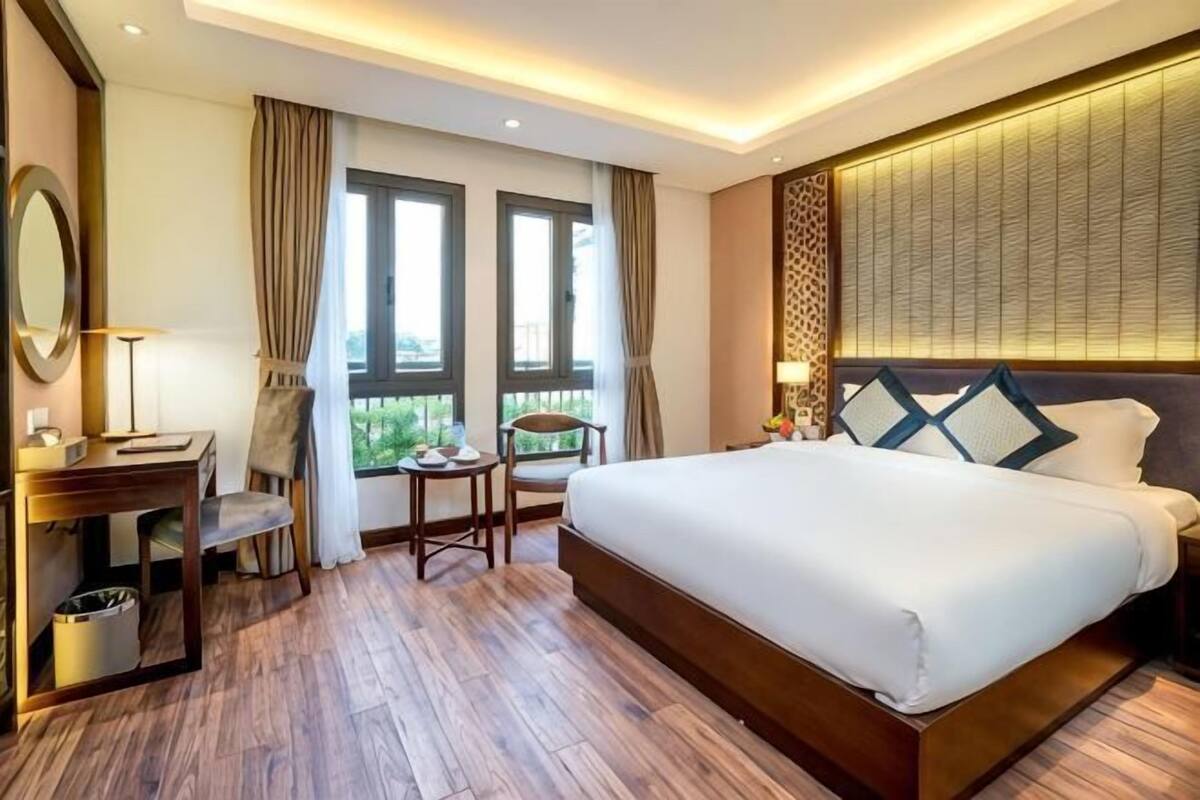 A hotel room with a large bed, a desk with a chair, a small table, and a window with curtains.