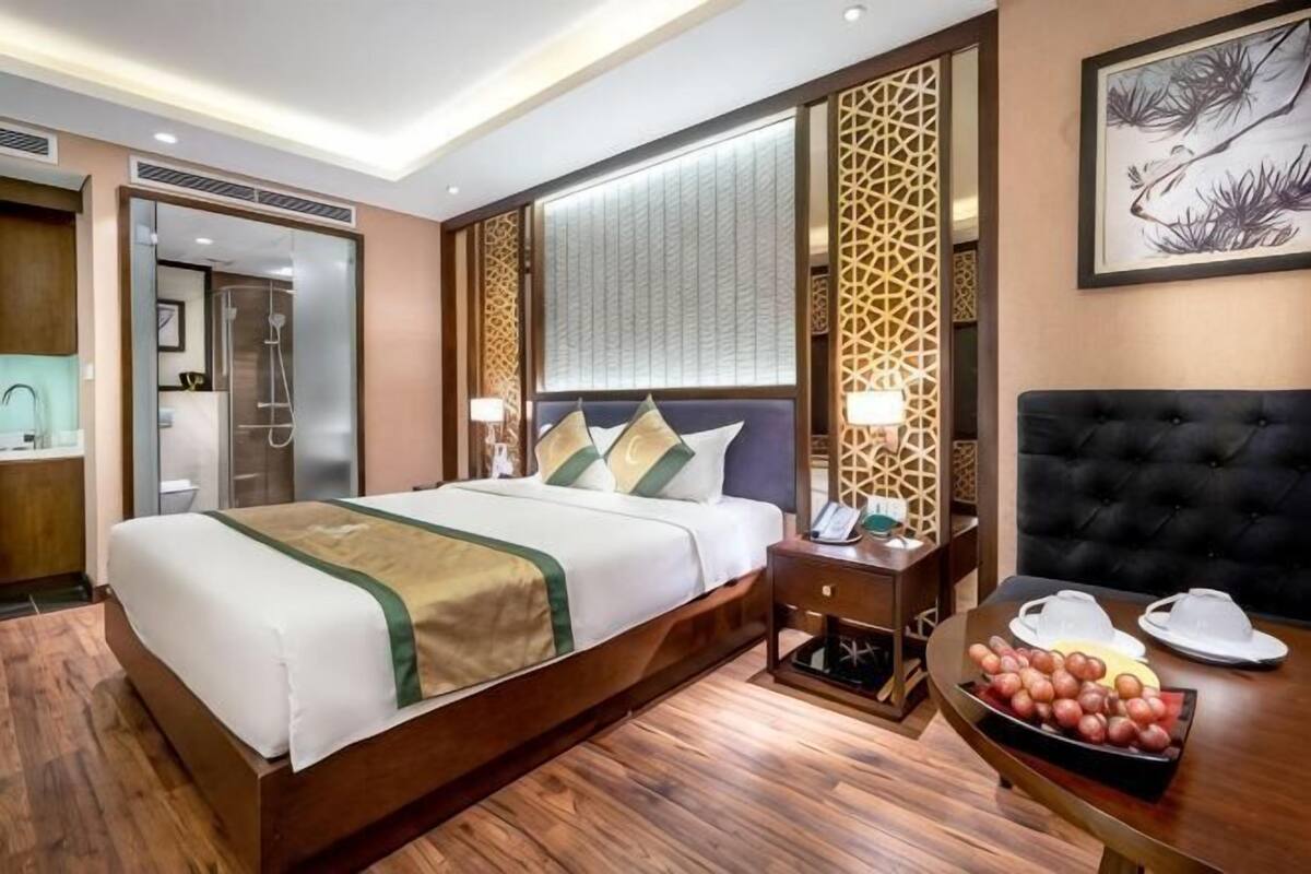 A hotel room with a large bed, bedside table, and a small dining table with fruit.