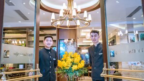 Two individuals standing in a hotel lobby with a chandelier, a large vase of yellow flowers, and a reception desk. - Conifer Grand Hotel (Hanoi)