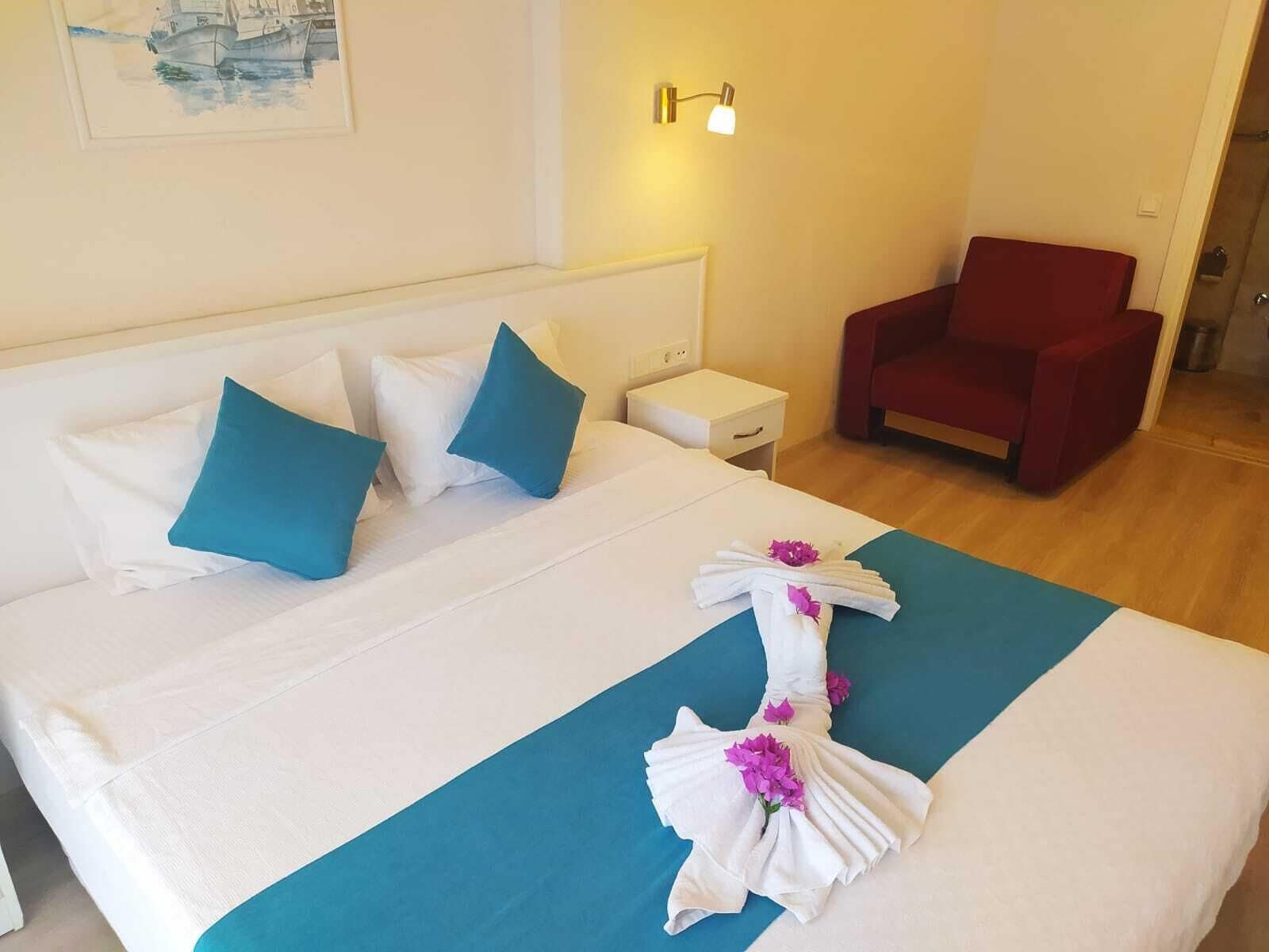 A neatly made bed with white and blue pillows, a teal bedspread, and a towel with flowers. There is a red chair and a small bedside table with a lamp.