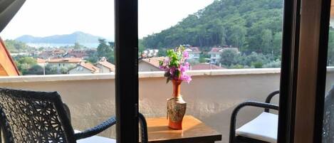 A balcony with wicker chairs and a table, a vase with flowers, and a view of houses and hills.