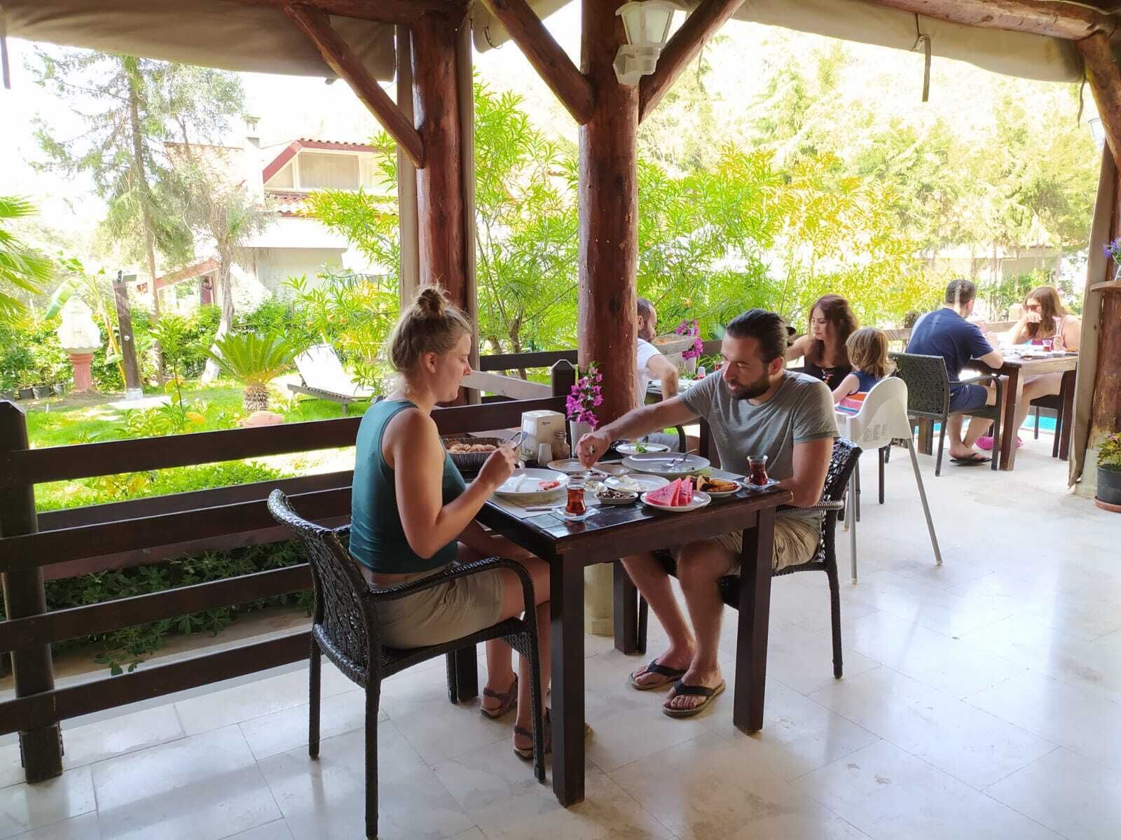 Un espace de restauration extérieur abrité, doté de tables et de chaises en bois, entouré de verdure et offrant une vue sur un quartier résidentiel.