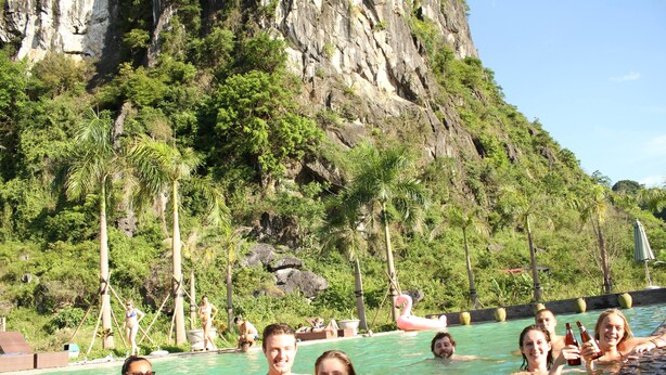 Una piscina con gente disfrutando en el agua, rodeada de vegetación exuberante y un acantilado rocoso.