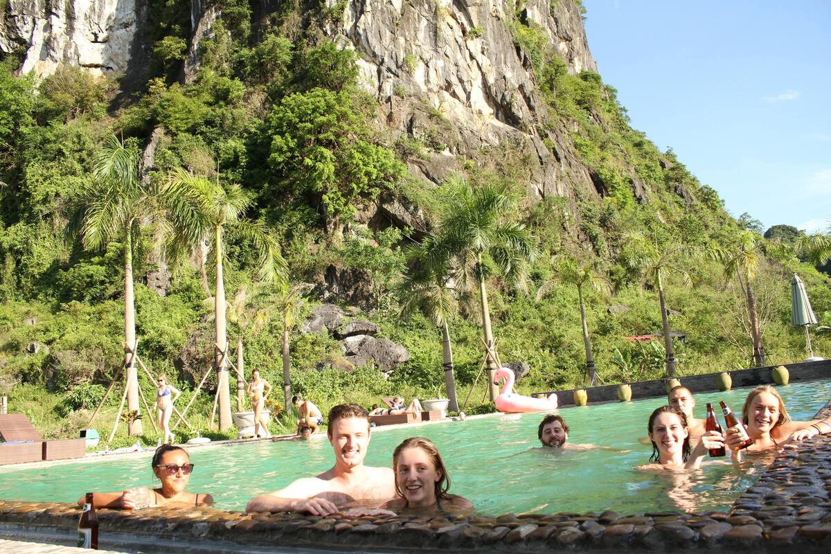 Piscina com pessoas aproveitando a água, rodeada por vegetação exuberante e um penhasco rochoso.