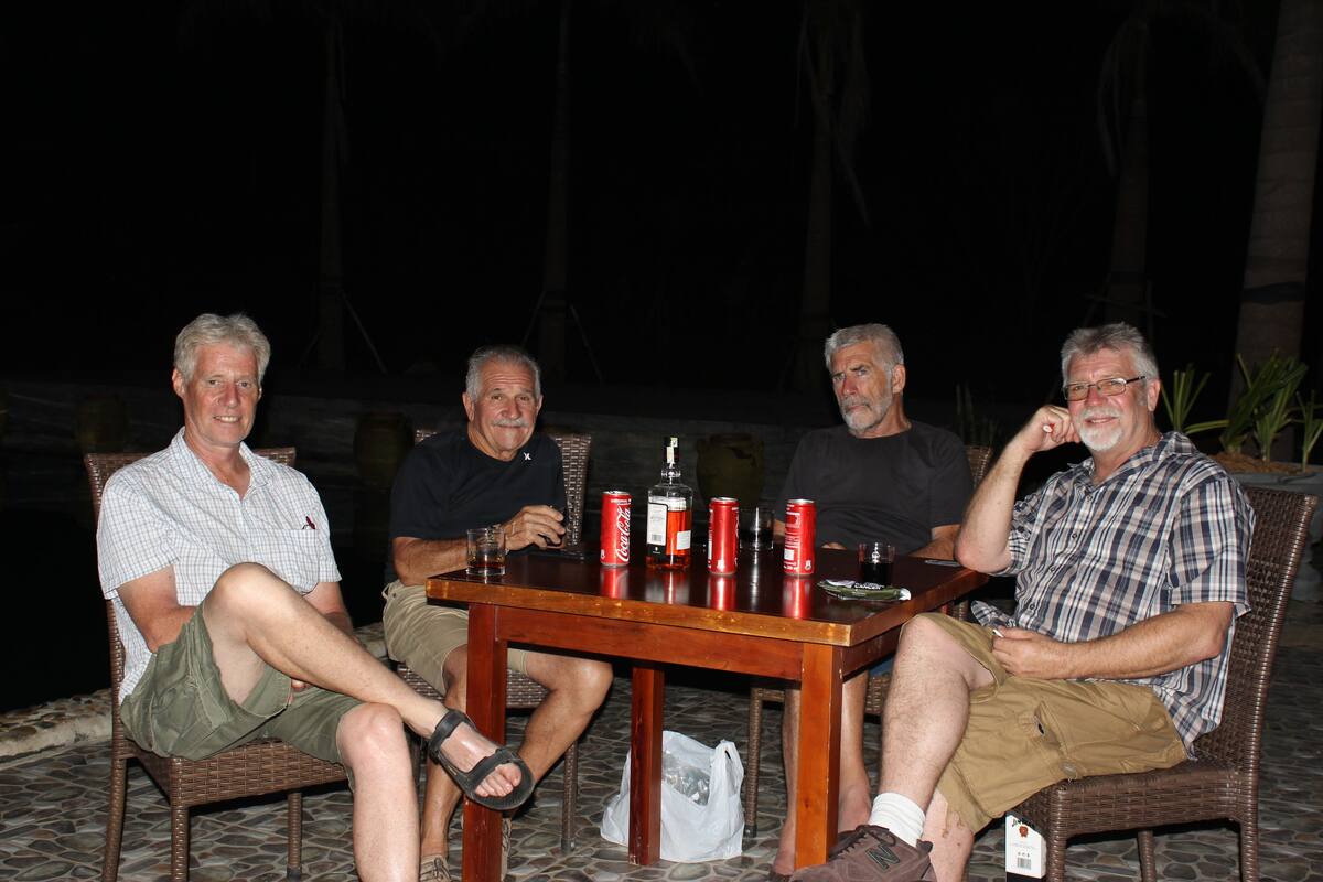 Quatro homens sentados em volta de uma mesa de madeira com bebidas e latas, ao ar livre à noite.