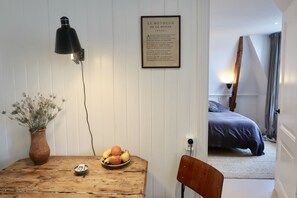 Een kamer met een houten tafel, een vaas met bloemen, een schaal fruit, een gedicht in een lijst aan de muur, en een bed op de achtergrond.