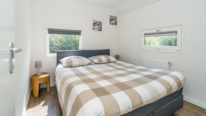 A bedroom with a bed, two windows, a radiator, and a small wooden stool.