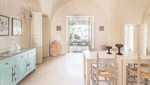 En rummelig stue med et højt hvælvet loft, spiseplads med bord og stole, og en stor vase på et skab.