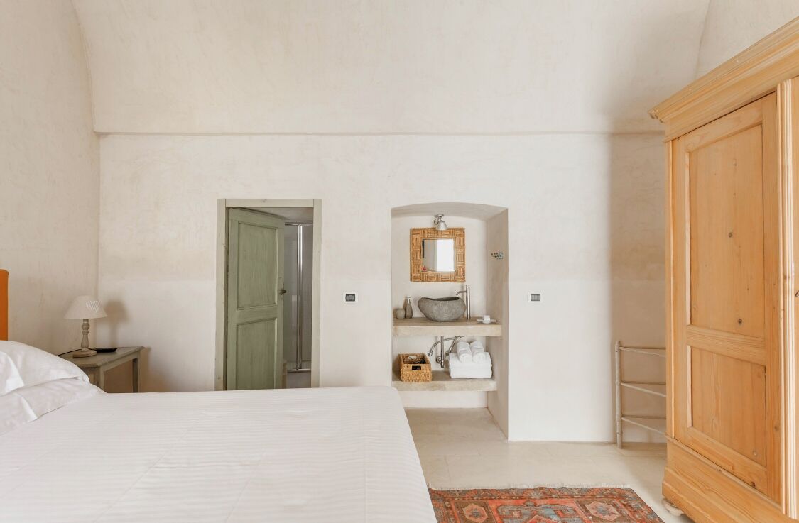 Un dormitorio con cama, un armario de madera, un baño con lavamanos y espejo, y una puerta que da a otra habitación.