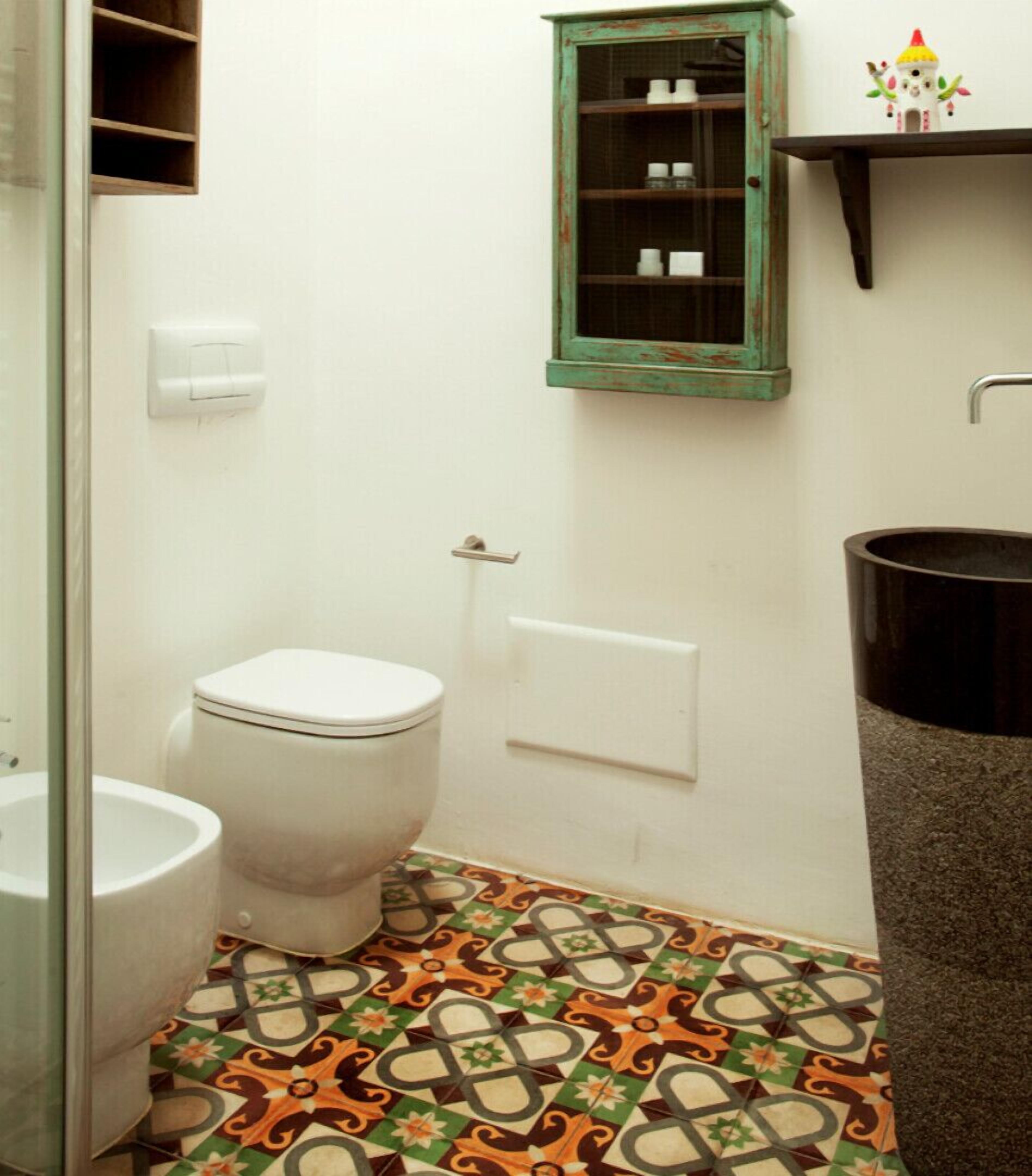 Un baño con inodoro, lavamanos, cubo de basura, un armario con puertas de vidrio y baldosas de suelo estampadas.