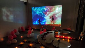 A room with a large screen displaying a space-themed projection, a table with candles and a decorative arrangement, and balloons scattered around.