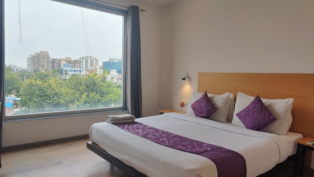 A hotel room with a large window, a bed with white bedding and purple pillows, a wooden headboard, and a nightstand with a lamp.